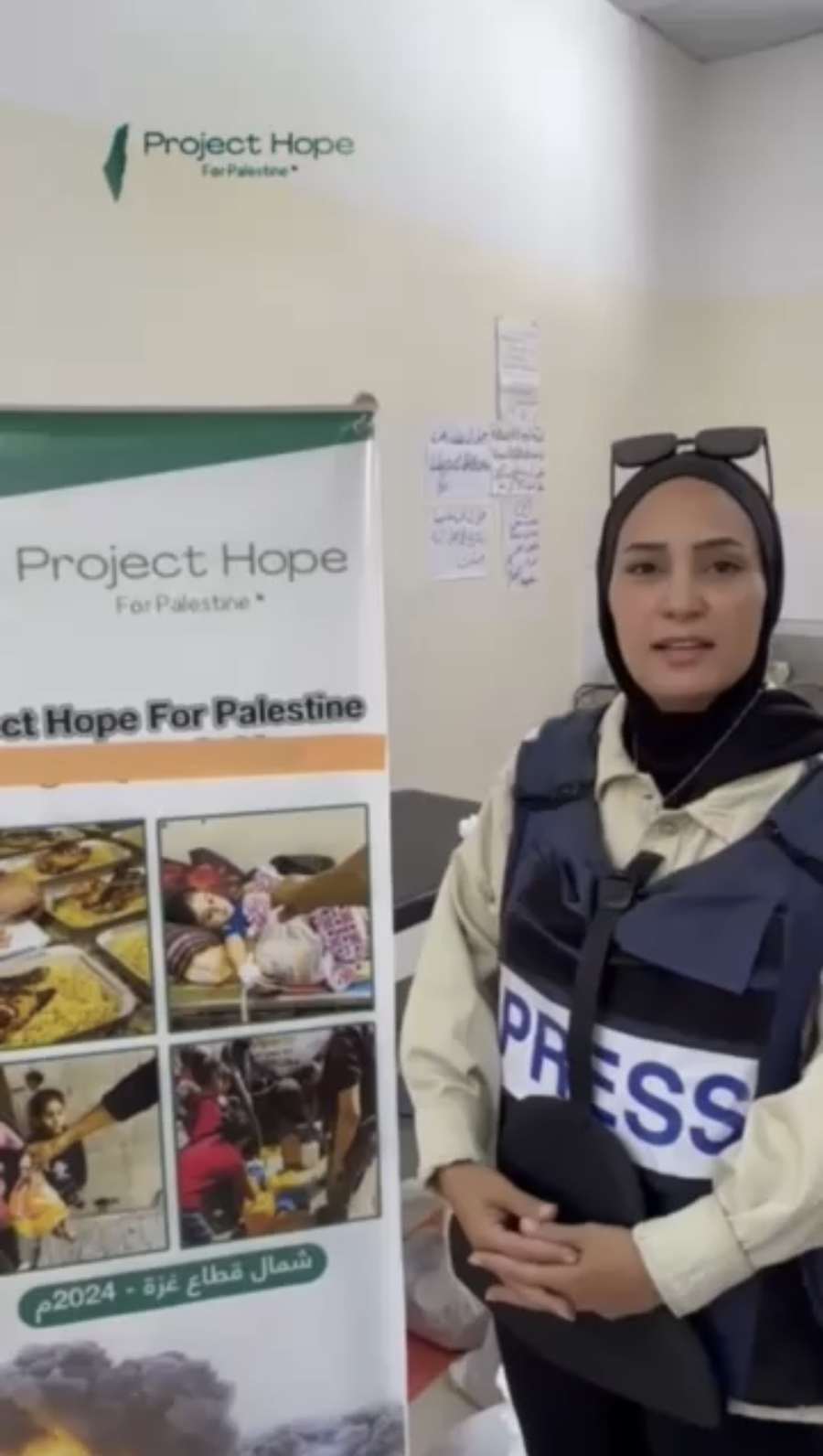 A Palestinian journalist in a press vest beside a Project Hope banner in north Gaza